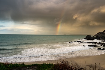 Strip of beach flanked by rocks on the ocean with rainbow on the horizon. 