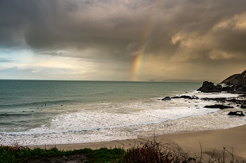 Strip of beach flanked by rocks on the ocean with rainbow on the horizon.