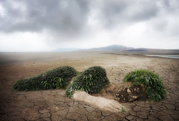 An andorgenous figure scuplted from mud lies on dry, cracked lake bed with mountains in the distance.